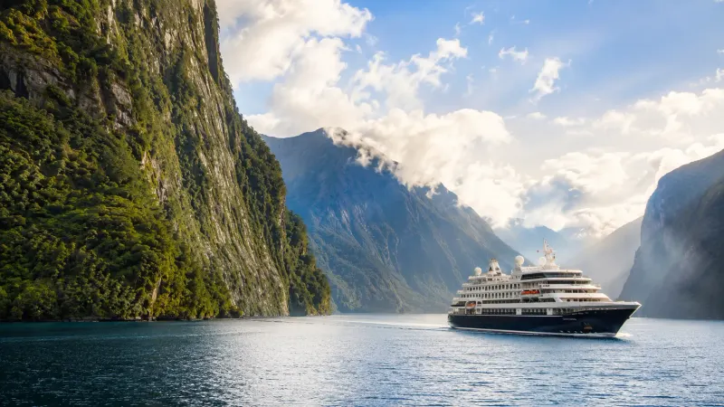 A cruise vessel navigating the dramatic mountain-bordered waters of Milford Sound fjord in New Zealand, a cruise organized by Piccola Travel, a Miami travel agency.
