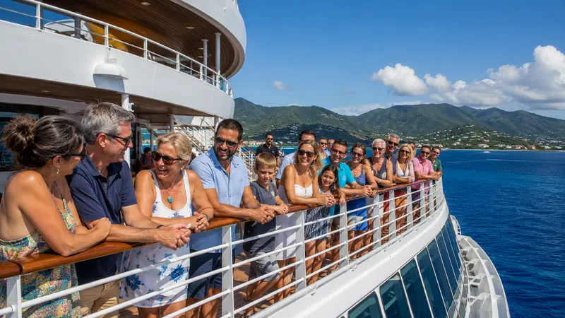 Family and friends gathered along a cruise ship deck railing overlooking a tropical coastline on a group cruise booked through Piccola Travel, a Miami travel agency.