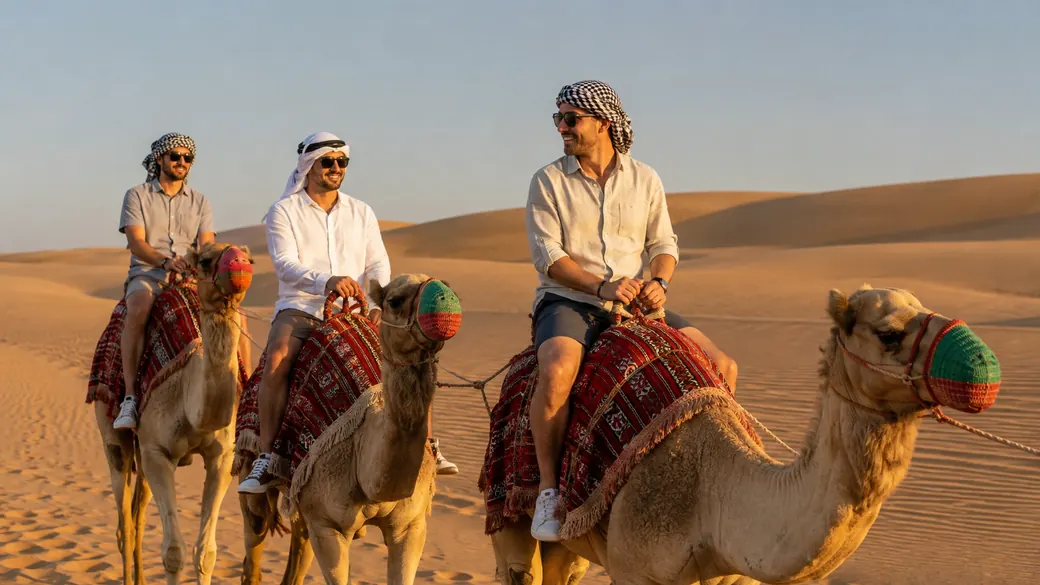 Three travelers in traditional headdresses ride camels across golden desert dunes on a Dubai desert safari tour