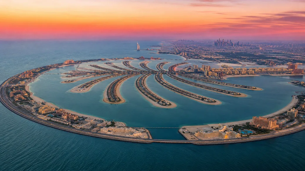 Aerial view of the Palm Jumeirah island and turquoise coastline at sunset with the sail-shaped hotel on the horizon
